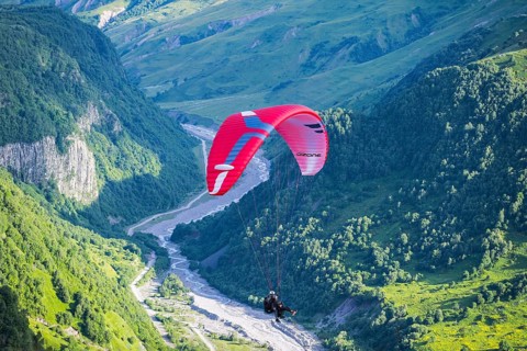 A paraglider floats over a narrow, green valley region with a fast-flowing river, paraglider near