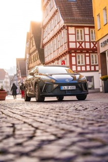 Car drives through an old town with half-timbered houses, some people on foot, MG4 electric car,