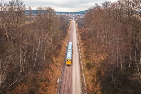 Long view of a rail line with a yellow train through a wintery forest, using a tamping machine at