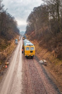 Train with construction workers on a rail line surrounded by winter forest, using a tamping machine