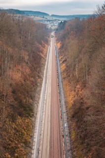 A long railway line leads through a hilly autumn landscape with bare trees, using a tamping machine