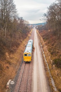 Yellow train during construction on a long rail line surrounded by bare trees, using a tamping