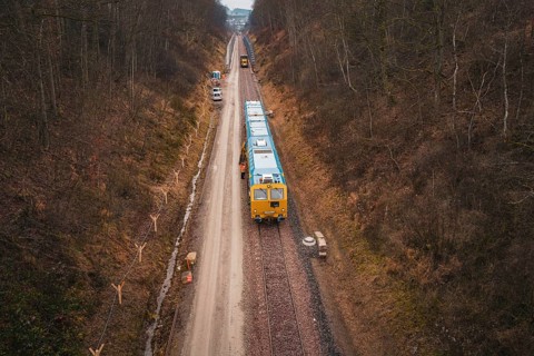 View of a railway line with a yellow train in a lightly wooded area, tamping machine used at the