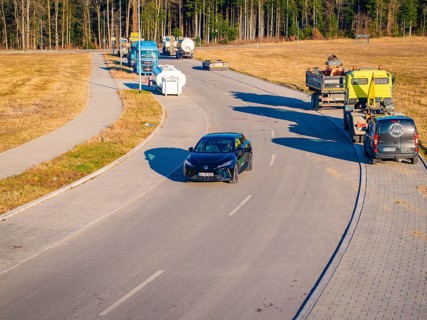 A car drives on a wide road surrounded by trucks and forest in the background, MG4 electric car,