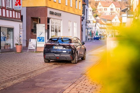 A car drives through a charming shopping street with half-timbered houses and blooming flowers, MG4