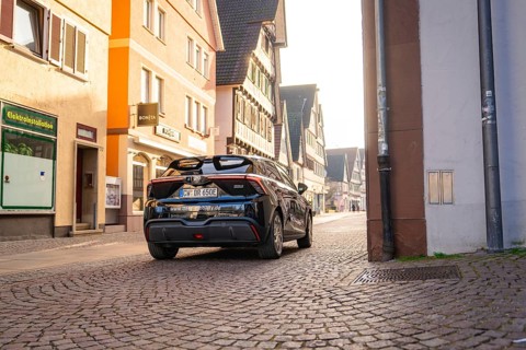 Black car drives through a sunny street lined with historic buildings, MG4 electric car, Deer