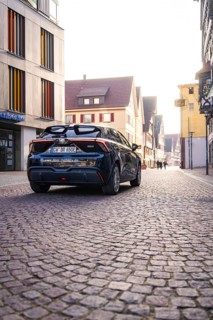 Black car drives through a paved city street between modern and historic buildings in sunlight, MG4