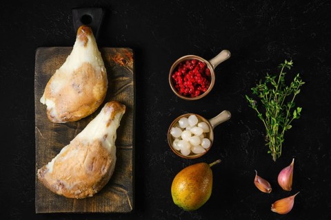Two large duck confit covered with congealed fat on wooden board next to pear, lingonberry, garlic