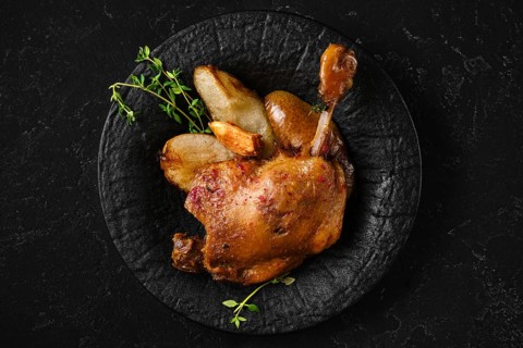 Top view of roasted duck leg on a textured black plate with caramelised in duck fat slices of pear.