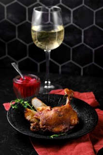 Duck confit with roasted pears on a black plate next to fruit jam and a glass of white wine. The