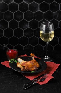 A plate with duck confit on a red cloth served with a glass of white wine and a lingonberry sauce