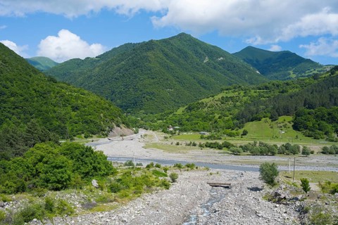 A riverbed leads through green mountains under a partly cloudy, blue sky, landscape near Chartali,