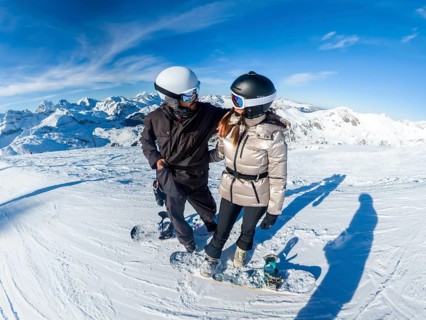 Couple preparing to snowboard on a sunny mountain peak, wearing helmets and goggles with snow