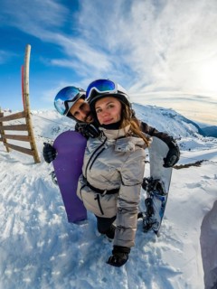 Young couple in helmets and goggles smile at camera, embracing with snowboards on a bright sunny