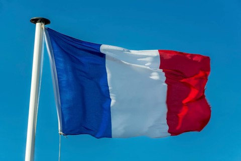 A French flag flutters in the wind on a white flagpole. The background is a clear, azure sky
