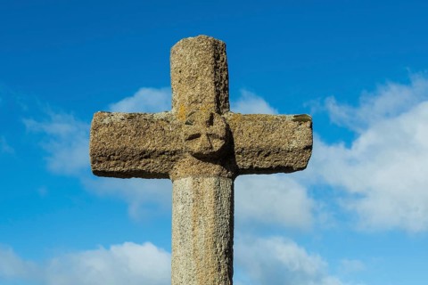 Stone cross stands tall against a bright blue sky with scattered white clouds in the background,