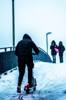 Winter weather, blowing snow, man with e-scooter trying to make his way uphill on a snowy sidewalk,