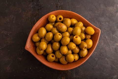 Fresh green olives in ceramic clay bowl on black concrete background. Top view, flat lay, close up