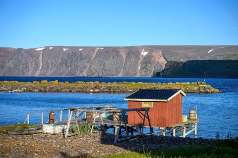 Small wooden cabin on a calm blue sea with mountain backdrop, red house on the shore with a view of
