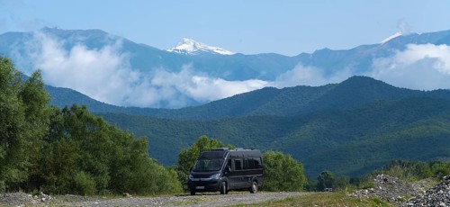 A black van on a road in a mountain valley with snowy peaks and clouds, camper in front of the