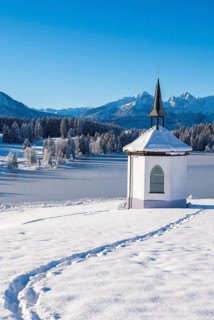 A small chapel in a snow-covered landscape with traces in the snow and mountains in the background,