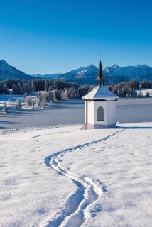 A small chapel in a snowy landscape with mountains in the background and a clear blue sky,