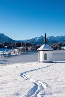 A small chapel stands on a snowy meadow against a mountain backdrop under clear, blue sky,