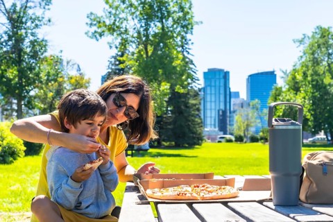 Mother helps her son eat a slice of pizza at a picnic table in a calgary park, enjoying quality