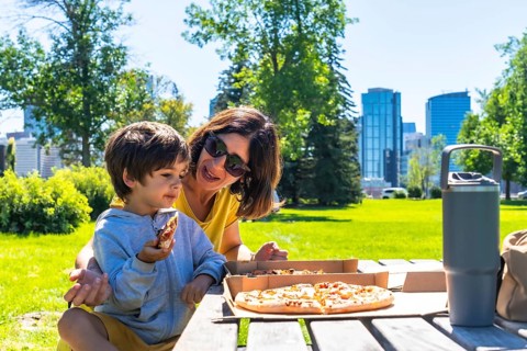 Mother and son are sharing a pizza picnic lunch together on a sunny day at a park in calgary,