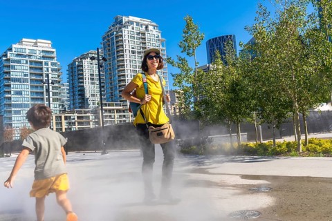 Tourist in a yellow shirt and backpack smiling while child runs through a refreshing misting system