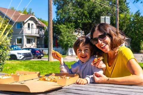 Happy mother and son laughing and eating pizza outdoors at a picnic table in the suburbs of