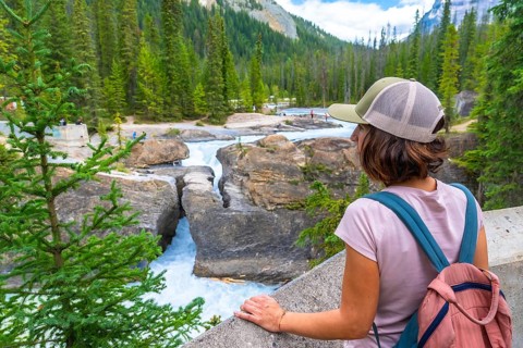 Female tourist wearing a baseball cap and backpack, admiring the bow river natural bridge in banff