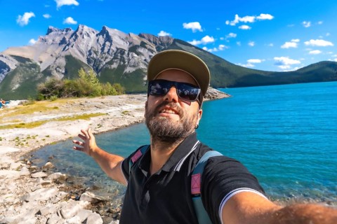Happy tourist taking a selfie with outstretched arm by turquoise lake minnewanka, surrounded by the