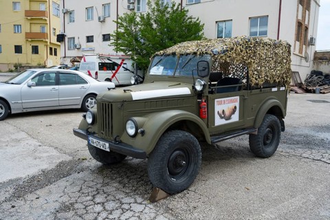 A green vintage car with a camouflage net is parked in a parking lot in front of a building, Soviet