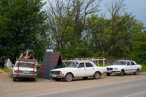 Three older white cars are parked on a rural road next to trees under a cloudy sky, cars with roof