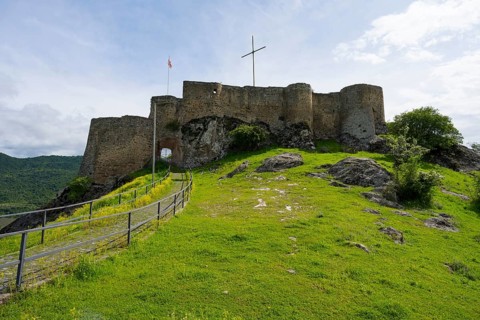 The picture shows medieval castle walls on a hill with surrounding nature under a cloudy sky,