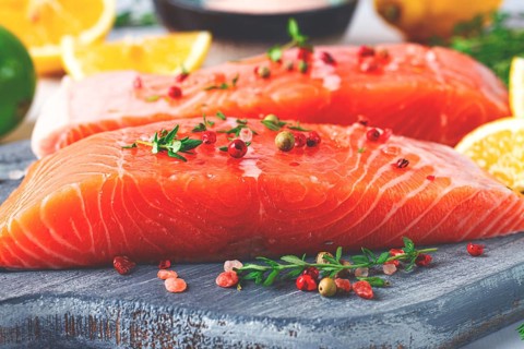 Two pieces of salmon fillet sit on a wooden cutting board. They are topped with herbs and pink