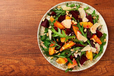 A bowl of salad contains beetroot, orange slices, pine nuts, and leaves. It is placed on a wooden