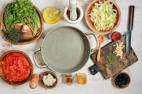Various food items and cooking tools are arranged on a wooden table. Herbs, vegetables, and spices