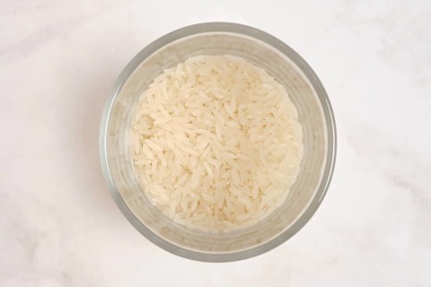 A clear bowl contains uncooked white rice. The rice is fluffy and dry, waiting to be cooked. The