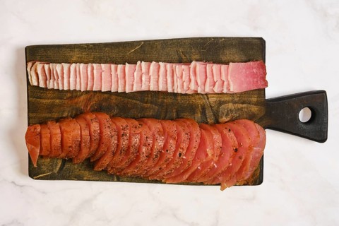 A wooden cutting board displays two types of cured meat. One side has thin slices of pink meat