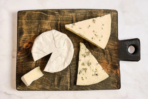 A wooden board holds pieces of cheese cut into various shapes. One piece is partially sliced,
