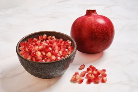 A bowl contains bright pomegranate seeds next to a whole pomegranate. The bowl is placed on a light
