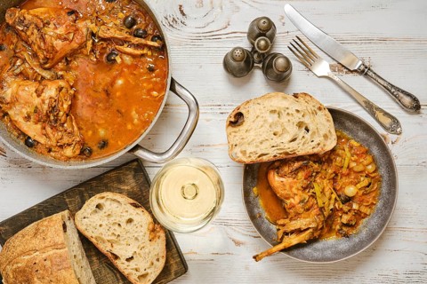 A rabbit stew with olives and vegetables sits in a pan next to a bowl. A piece of bread lies beside