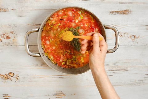 A hand sprinkles spice in a pot with chopped vegetables and a rich tomato base. Spices are added to