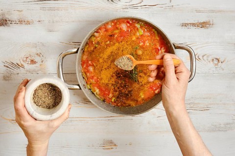 A person adds pepper to a pot of vegetables and tomatoes. The hands hold a wooden spoon and a