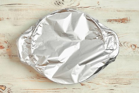 A roasting pot is covered with aluminum foil placed on a wooden kitchen table. The foil prevents