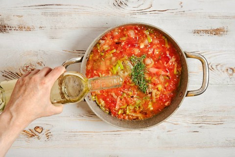 A person pours white wine into a pot filled with chopped tomatoes and vegetables. The pot sits on a
