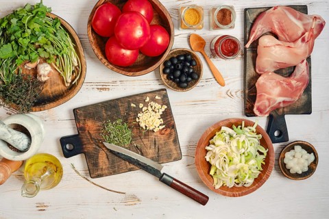 Fresh tomatoes, herbs, and meat are spread on a wooden table. There are bowls of spices and chopped