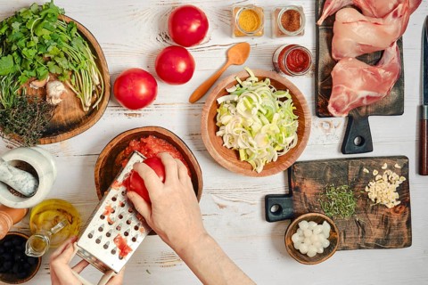 A person is grating tomatoes and chopping vegetables. Fresh herbs, spices, and rabbit meat are laid